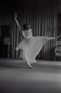 A person is gracefully dancing in a flowing dress, captured in mid-motion with one leg extended and arms raised. The backdrop consists of elegant curtains and a partially visible chair on the right side. The scene appears to be indoors, with subtle lighting accentuating the movement.