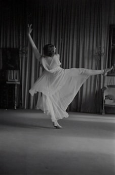 A person is gracefully dancing in a flowing dress, captured in mid-motion with one leg extended and arms raised. The backdrop consists of elegant curtains and a partially visible chair on the right side. The scene appears to be indoors, with subtle lighting accentuating the movement.