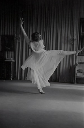 A person is gracefully dancing in a flowing dress, captured in mid-motion with one leg extended and arms raised. The backdrop consists of elegant curtains and a partially visible chair on the right side. The scene appears to be indoors, with subtle lighting accentuating the movement.