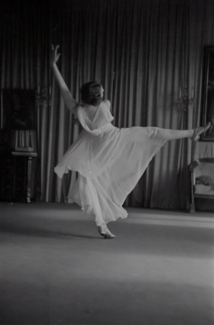 A person is gracefully dancing in a flowing dress, captured in mid-motion with one leg extended and arms raised. The backdrop consists of elegant curtains and a partially visible chair on the right side. The scene appears to be indoors, with subtle lighting accentuating the movement.