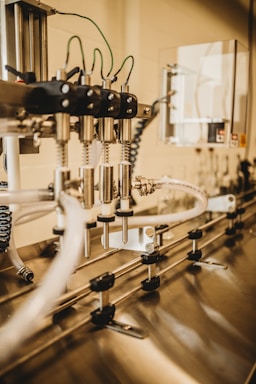 A close-up view of an industrial machine component featuring multiple metal pipes and mechanical parts. Various cables are connected, and the setting appears to be a factory or manufacturing environment. The image has a warm, metallic look with reflections off the metal surfaces.
