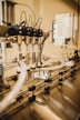 A close-up view of an industrial machine component featuring multiple metal pipes and mechanical parts. Various cables are connected, and the setting appears to be a factory or manufacturing environment. The image has a warm, metallic look with reflections off the metal surfaces.