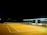 A tennis court is illuminated under bright floodlights at night. The court is empty, with no players present. The net is visible in the center of the court, and to the right, there are white benches and an umpire's chair. The surrounding area is dark, contrasting with the well-lit court.