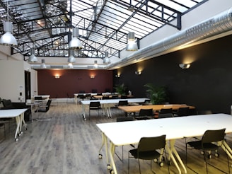 A spacious open-plan room with large skylights and industrial-style hanging lights. The room features white tables on wheels, black chairs, and modern decor. The floor is wooden, and the walls have a dark color with wall-mounted light fixtures. There are plants in the room, adding a touch of greenery.