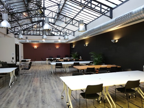 A spacious open-plan room with large skylights and industrial-style hanging lights. The room features white tables on wheels, black chairs, and modern decor. The floor is wooden, and the walls have a dark color with wall-mounted light fixtures. There are plants in the room, adding a touch of greenery.