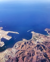 Aerial view of a coastal region featuring a harbor with docks and ships. The landscape includes rugged mountains and urban settlements adjacent to expansive blue water. The coastline wraps around the water with visible roads connecting the areas. A boat is navigating through the water, creating a white wake.