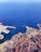 Aerial view of a coastal region featuring a harbor with docks and ships. The landscape includes rugged mountains and urban settlements adjacent to expansive blue water. The coastline wraps around the water with visible roads connecting the areas. A boat is navigating through the water, creating a white wake.