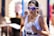 A person is running while wearing a white athletic tank top and reflective sunglasses. They are holding a packet of gel or energy drink in one hand. The background is blurred, suggesting motion and focus on the subject.