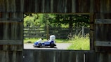 A framed view through a wooden structure shows a small blue and white go-kart on a paved path. In the background, there is a wooden fence and lush green foliage, suggesting a natural, outdoor setting.