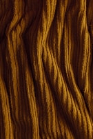 Ribbed fabric displaying vertical, textured patterns in a warm, golden-brown color. The surface appears soft and slightly folded, suggesting a material like corduroy or velvet.