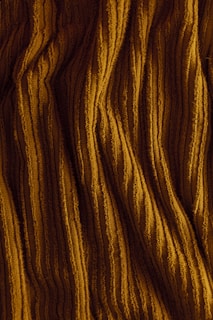 Ribbed fabric displaying vertical, textured patterns in a warm, golden-brown color. The surface appears soft and slightly folded, suggesting a material like corduroy or velvet.