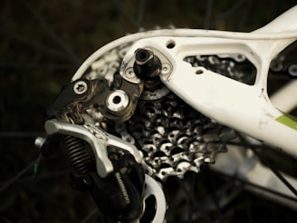 Close-up of a bicycle's rear derailleur and gear cassette. The metallic components contrast with the white and slightly worn bicycle frame, featuring intricate details and mechanical parts including the chain and bolts.