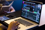 A person is using a laptop displaying audio mixing software with various faders, graphs, and control panels on the screen. Another individual is sitting nearby, holding and reading a book. There are additional devices on the table, including a smartphone and possibly a tablet, with one also showing a digital interface.