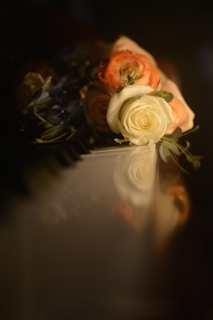 A bouquet of roses, featuring white and peach-colored flowers, lies on a reflective surface, possibly a piano. Soft lighting creates a warm and serene atmosphere, highlighting the delicate petals and subtle shadows.