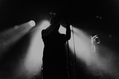 A silhouette of a person holding a microphone, standing in front of bright stage lights. The scene is set in a dimly lit environment, creating a dramatic contrast between the dark figure and the illuminated background.
