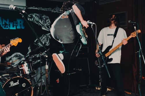 The image captures a live music performance with musicians in action on stage. One musician is energetically jumping while playing the electric guitar. The scene is dynamic and filled with musical equipment like microphones, drums, and an amplifier.