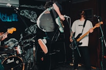 The image captures a live music performance with musicians in action on stage. One musician is energetically jumping while playing the electric guitar. The scene is dynamic and filled with musical equipment like microphones, drums, and an amplifier.