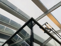 A modern architectural structure featuring a trapezoidal glass roof with several panels fitted with solar cells. The beams holding the glass panels are metallic, creating a sleek and industrial appearance. The perspective gives a sense of looking upwards through the roof, emphasizing the clean lines and geometric shapes.
