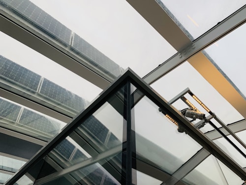 A modern architectural structure featuring a trapezoidal glass roof with several panels fitted with solar cells. The beams holding the glass panels are metallic, creating a sleek and industrial appearance. The perspective gives a sense of looking upwards through the roof, emphasizing the clean lines and geometric shapes.