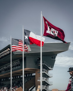 Three flags on poles are waving prominently against a gray sky. The flags include the United States, Texas, and a maroon flag with 'A&M' lettering. In the background, a large building with modern architecture features multiple levels and staircases. A person wearing maroon is visible in the foreground.
