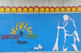 A mural features a stylized illustration of a person sweeping, along with vibrant sun motifs above the head and text in an Indian script. The artwork is predominantly blue and includes elements of orange, yellow, and green.