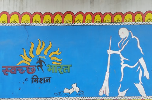 A mural features a stylized illustration of a person sweeping, along with vibrant sun motifs above the head and text in an Indian script. The artwork is predominantly blue and includes elements of orange, yellow, and green.