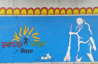 A mural features a stylized illustration of a person sweeping, along with vibrant sun motifs above the head and text in an Indian script. The artwork is predominantly blue and includes elements of orange, yellow, and green.