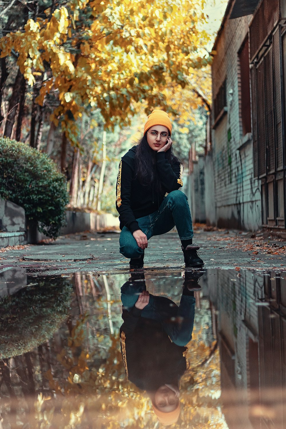 woman in black jacket, yellow beanie hat, and blue jeans kneeling on ground