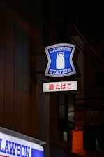A brightly illuminated sign with the words 'LAWSON STATION' and a graphic of a milk can is mounted on the side of a building at night. Below it is another sign with Japanese characters. The surrounding area is dimly lit, highlighting the blue and white colors of the signage.