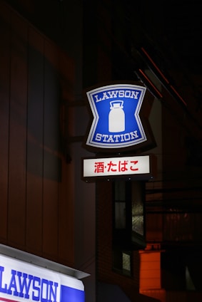 A brightly illuminated sign with the words 'LAWSON STATION' and a graphic of a milk can is mounted on the side of a building at night. Below it is another sign with Japanese characters. The surrounding area is dimly lit, highlighting the blue and white colors of the signage.