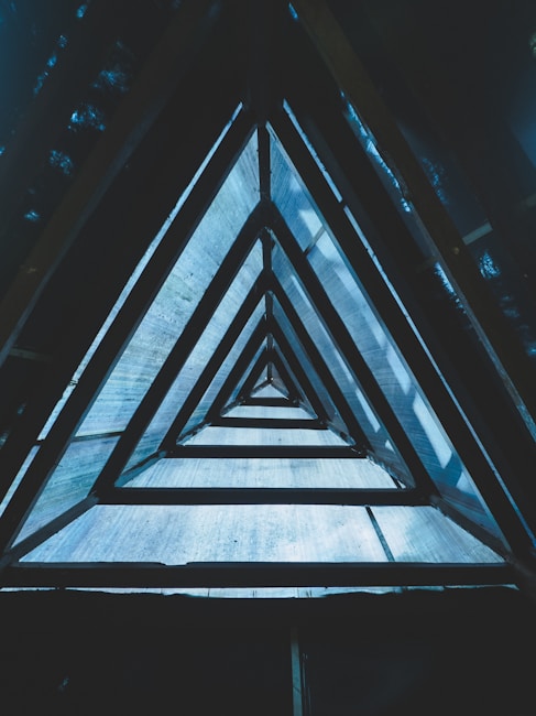 A geometric perspective showing a series of nested triangular frames, creating a tunnel-like effect. The triangles are made from a metallic material, and the scene is illuminated by soft blue lighting, casting shadows that accentuate the structure's depth.