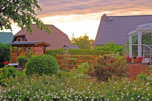 A vibrant garden featuring a variety of blooming flowers and well-maintained greenery, with colorful houses in the background. The setting sun casts a warm glow across the scene, highlighting a wooden gazebo and a glass conservatory. Potted plants and a neatly trimmed hedge add to the lush garden atmosphere.