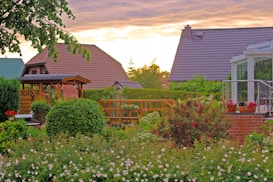 A vibrant garden featuring a variety of blooming flowers and well-maintained greenery, with colorful houses in the background. The setting sun casts a warm glow across the scene, highlighting a wooden gazebo and a glass conservatory. Potted plants and a neatly trimmed hedge add to the lush garden atmosphere.