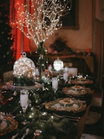 A festive dining table setting featuring an elegant arrangement with a central display of branches adorned with twinkling lights. The table is set with fine china, decorated with intricate golden details. Champagne flutes filled with a drink complement the setting, while additional sparkling decorations and greenery enhance the luxurious ambiance.
