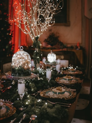A festive dining table setting featuring an elegant arrangement with a central display of branches adorned with twinkling lights. The table is set with fine china, decorated with intricate golden details. Champagne flutes filled with a drink complement the setting, while additional sparkling decorations and greenery enhance the luxurious ambiance.
