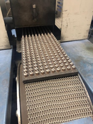 A factory conveyor belt with rows of small metal components arranged in a grid pattern. The components are evenly spaced on a textured mat, and the belt is moving through a metallic apparatus.