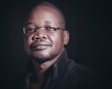 A man with short hair and glasses gazes confidently at the camera. He wears a dark, textured jacket and is set against a shadowy background.