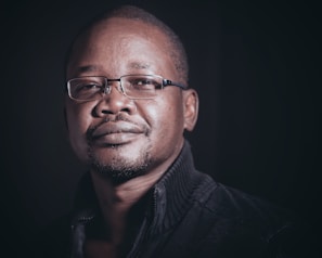 A man with short hair and glasses gazes confidently at the camera. He wears a dark, textured jacket and is set against a shadowy background.