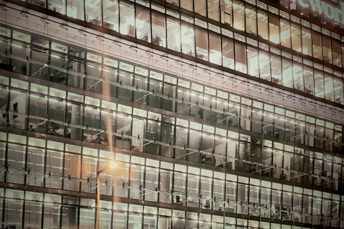 A multi-story office building with large glass windows showing illuminated office spaces at night. The interior lighting reveals office furniture and workstations inside, and the exterior reflects the glow of streetlights.