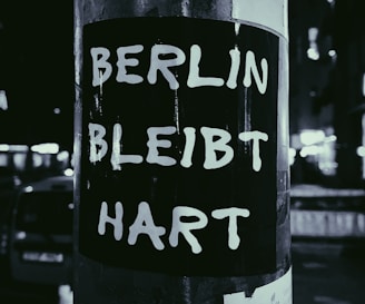 A black and white sticker with bold white text that reads BERLIN BLEIBT HART is affixed to a pole. The background features a blurred view of an urban street at night with lights and shadows.