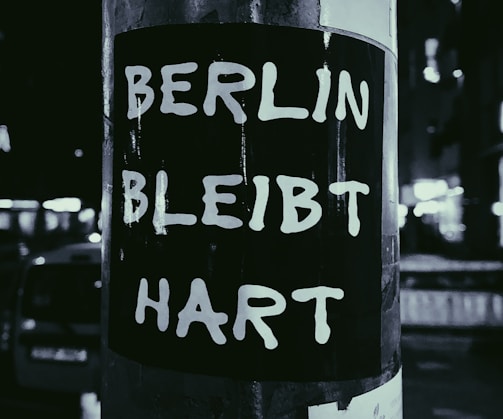 A black and white sticker with bold white text that reads BERLIN BLEIBT HART is affixed to a pole. The background features a blurred view of an urban street at night with lights and shadows.