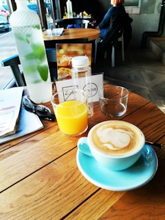 A cozy cafe setting with a wooden table holding a bottle of water with mint leaves, a glass of orange juice, and a cup of frothy cappuccino on a blue saucer. On the table, there are also a pair of sunglasses, a brochure, and a menu card. In the background, a person sits at another table near a window.