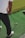 A person stands on a green artificial turf, holding a golf club, preparing to hit a yellow golf ball. The person is wearing a white shirt and dark pants with white and red shoes.