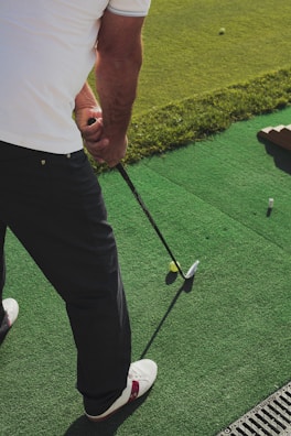 A person stands on a green artificial turf, holding a golf club, preparing to hit a yellow golf ball. The person is wearing a white shirt and dark pants with white and red shoes.