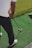 A person stands on a green artificial turf, holding a golf club, preparing to hit a yellow golf ball. The person is wearing a white shirt and dark pants with white and red shoes.