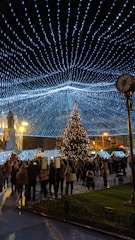People gather in a festive outdoor setting illuminated by a canopy of blue and white string lights. A decorated Christmas tree stands prominently in the center, surrounded by people walking and socializing. Buildings in the background have a warm glow, and there is a mix of light and shadow across the scene.