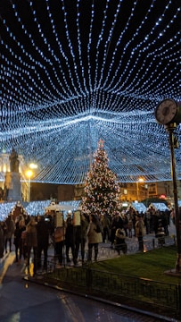 People gather in a festive outdoor setting illuminated by a canopy of blue and white string lights. A decorated Christmas tree stands prominently in the center, surrounded by people walking and socializing. Buildings in the background have a warm glow, and there is a mix of light and shadow across the scene.
