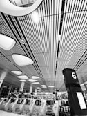 A modern indoor space featuring a high ceiling with linear patterns and futuristic lighting fixtures. Rows of kiosks and a pillar marked with a number 6 are visible, suggesting an organized and spacious environment. People are visible in the background beneath the structured design.
