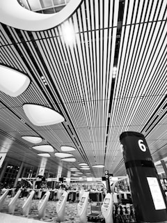 A modern indoor space featuring a high ceiling with linear patterns and futuristic lighting fixtures. Rows of kiosks and a pillar marked with a number 6 are visible, suggesting an organized and spacious environment. People are visible in the background beneath the structured design.