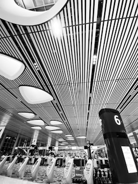 A modern indoor space featuring a high ceiling with linear patterns and futuristic lighting fixtures. Rows of kiosks and a pillar marked with a number 6 are visible, suggesting an organized and spacious environment. People are visible in the background beneath the structured design.